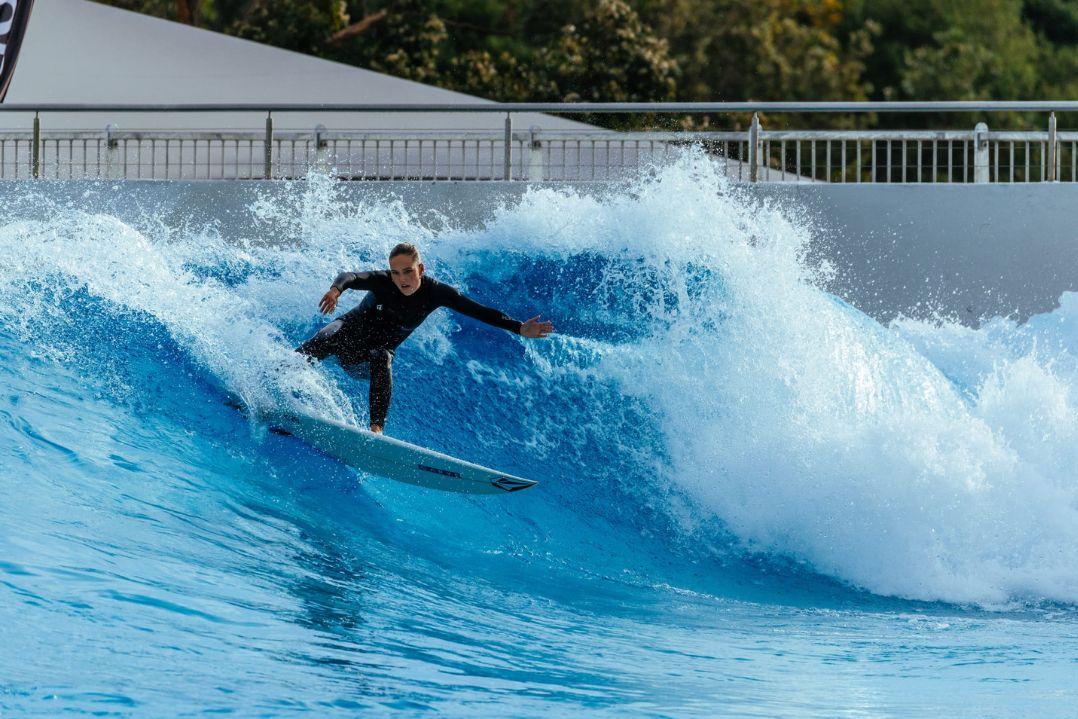 A surfer in a wetsuit skillfully rides a large, blue artificial wave in a wave pool. The scene conveys excitement and focus against the backdrop of trees.
