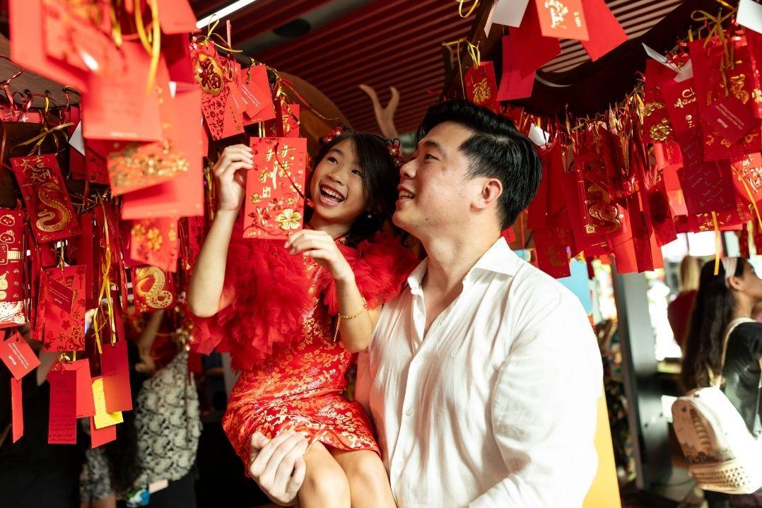 A couple joyfully interacts with vibrant red lanterns in a festive setting. The scene is lively and colourful, evoking a sense of celebration and happiness.