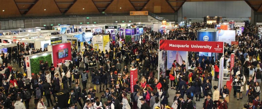 A bustling trade show with numerous booths in a large, arched venue. Crowds of people are engaging with exhibitors and exploring displays.
